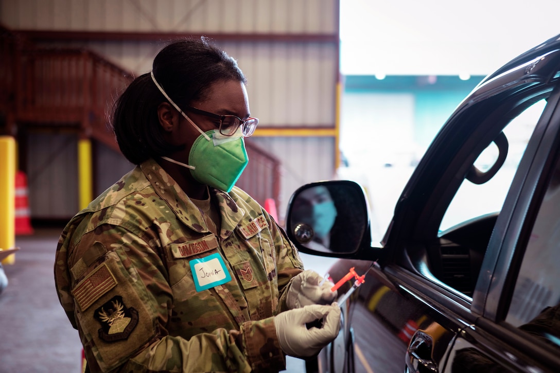 An airman wearing a face mask and glove holds a syringe before vaccinating someone in their vehicle.