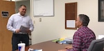 One man sits at his desk while speaking to a man who is standing.