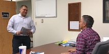 One man sits at his desk while speaking to a man who is standing.