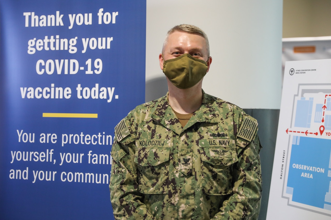 A sailor wearing a face mask poses in front of a COVID-19 vaccination banner.