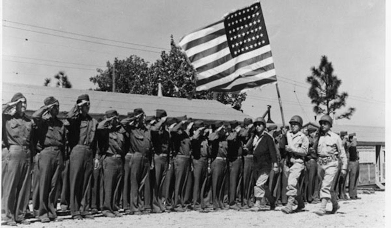 Men in uniform salute as three men walk by carrying a U.S. flag.