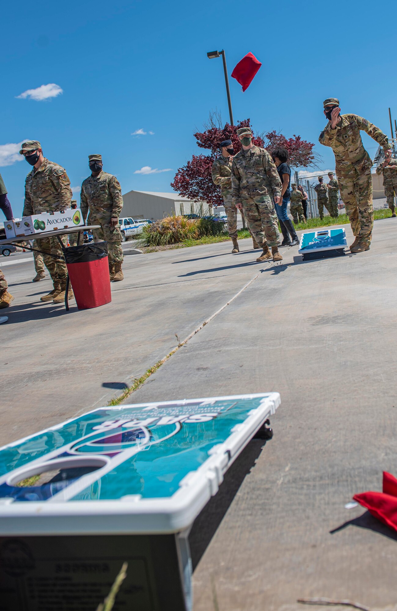 A male Airman has just tossed a cornhole bag and the bag is still mid-air before reaching the cornhole board at the other end of the photo.