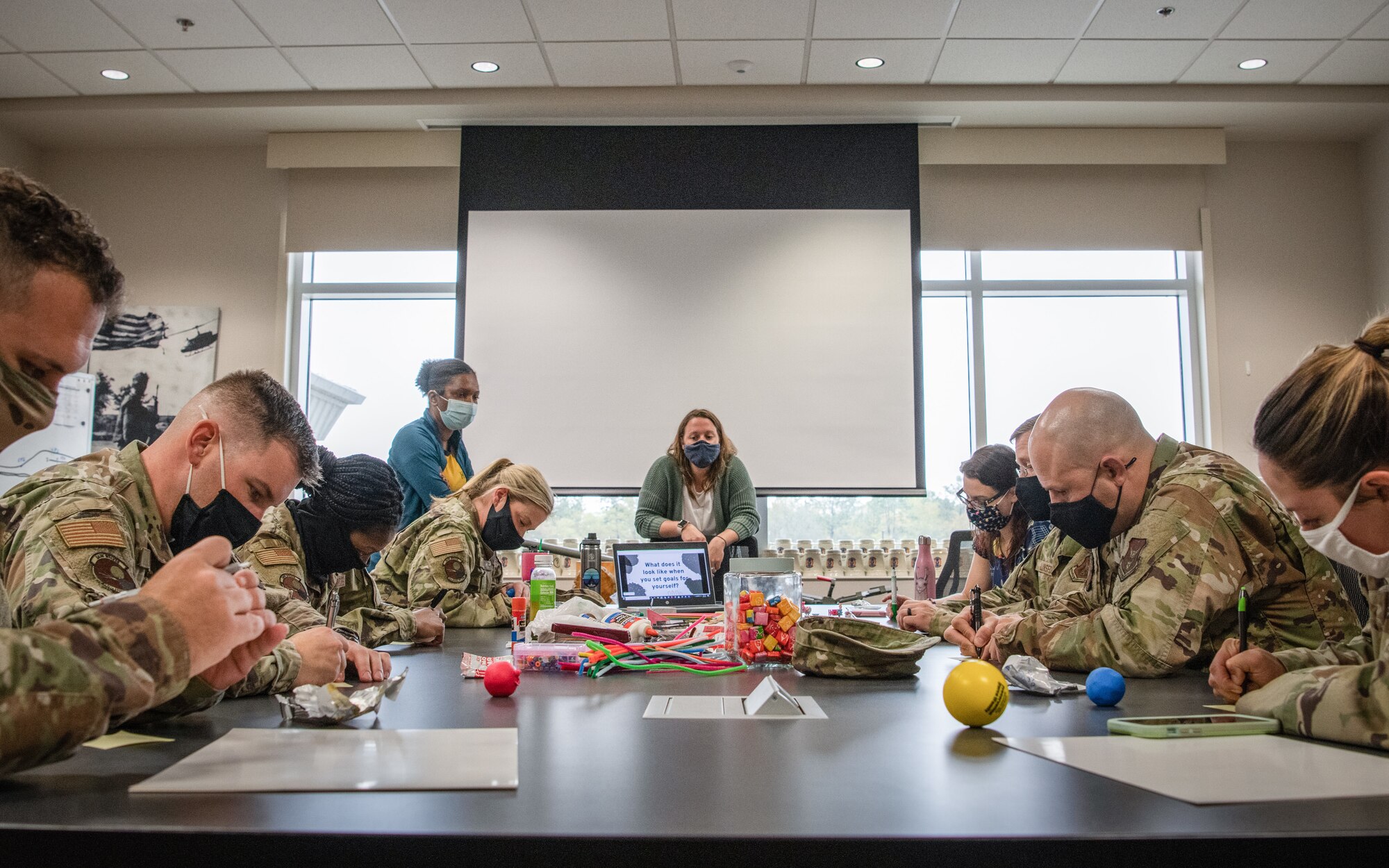 A group of Airmen sit at a table and write on notepads as a civilian woman watches.