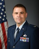Col. David Black poses in front of a gray field with and American flag behind his right shoulder.