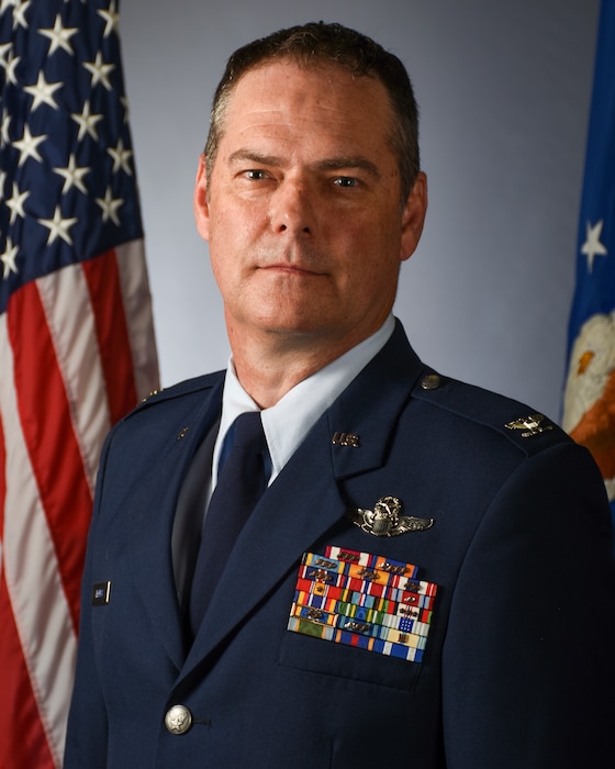 Colonel Michael Leonas poses in front of a gray field with an American flag behind his right shoulder and a US Air Force flag behind his left.