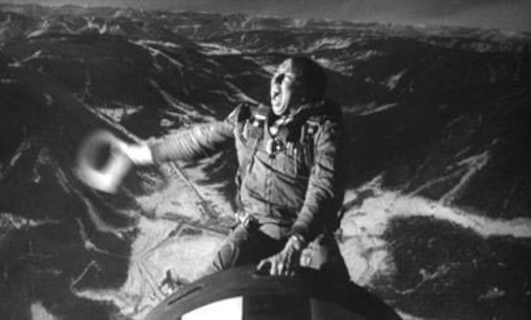 An actor, with a cowboy hat and dressed in a flight suit, rides a bomb.