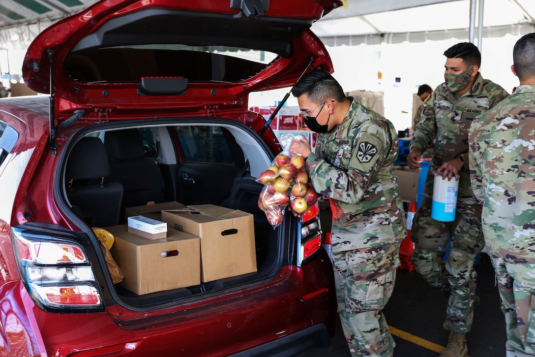 Three soldiers wearing face masks load groceries into the trunk of a vehicle.