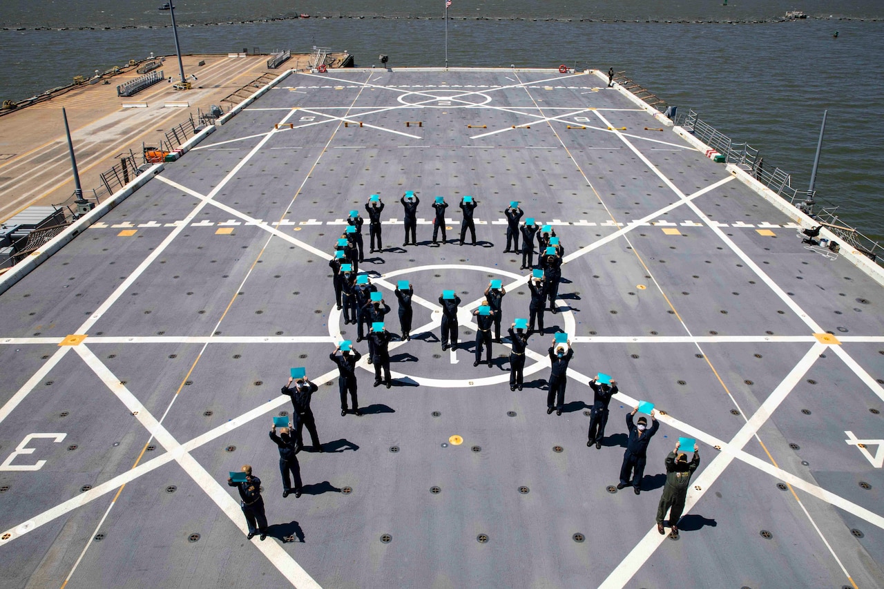 Sailors hold up pieces of teal paper while standing in a formation on the deck of a ship.