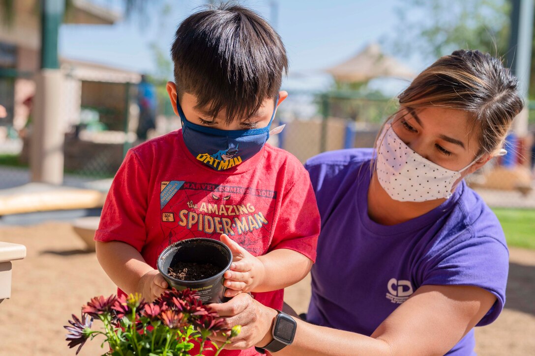 An adult helps a child pour soil into a flower pot.