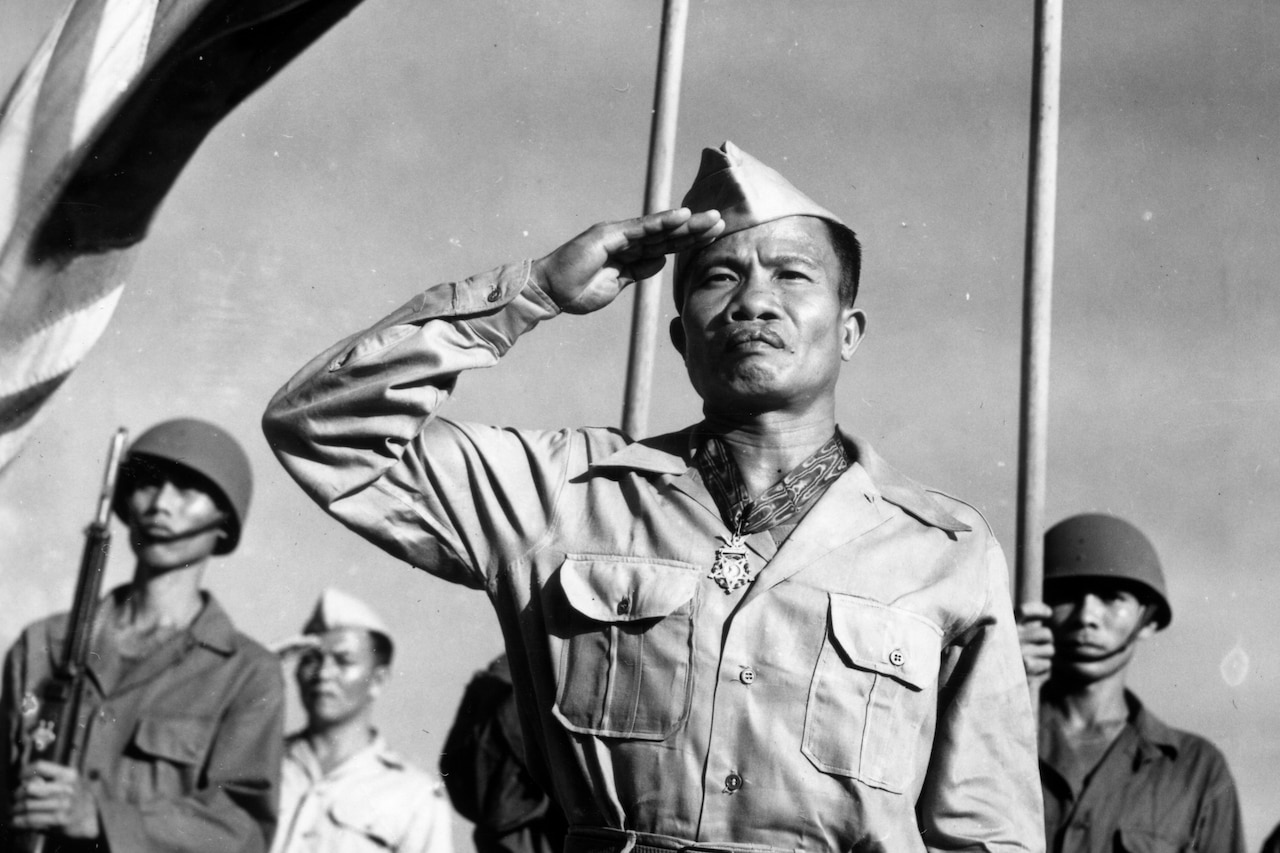 A soldier salutes while wearing a Medal of Honor, as fellow soldiers stand behind him outside and a flag waves above them.