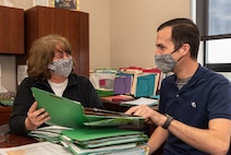 Photo of a woman is wearing a black top with a white design, while the man is wearing a dark blue shirt.  Behind them, you can see warehouse racks with boxes on them.