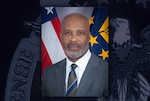 Head and shoulders portrait of a black man in dark suit and tie in front of the US flag.