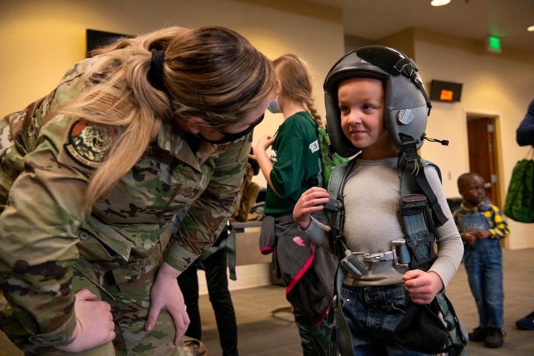 An airman talks to a military child wearing aircrew equipment.