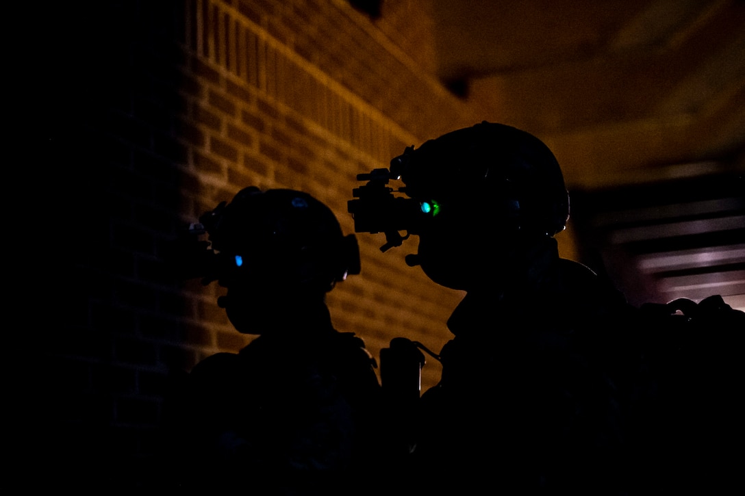 Two service members stand near a brick wall wearing helmets at night.