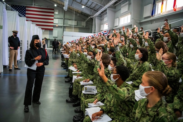 A Navy officer wearing a face mask speaks to new Navy recruits wearing face masks and seated on bleachers.