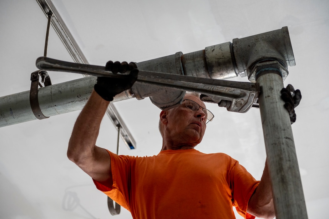A man in an orange shirt fitting a pipe with a large wrench.