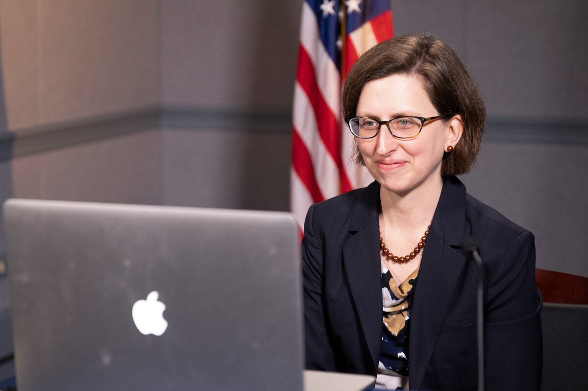 A woman looks at a laptop screen; a U.S. flag is in the background.