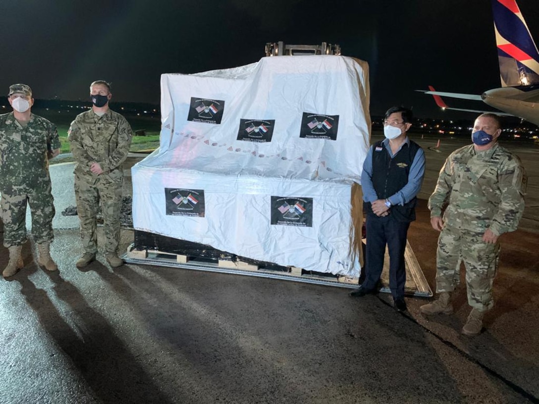 Three men in military uniforms and one in civilian clothes pose in front of a shipment of supplies covered with a protective sheet adorned with the flags of the U.S. and Paraguay.
