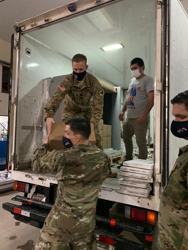 Three men in military uniforms and another in civilian clothes unload a refrigerated truck.