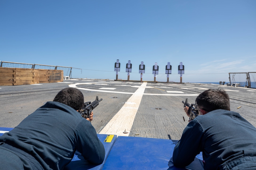 Two sailors aim their rifles at targets while lying on the deck of a military ship.