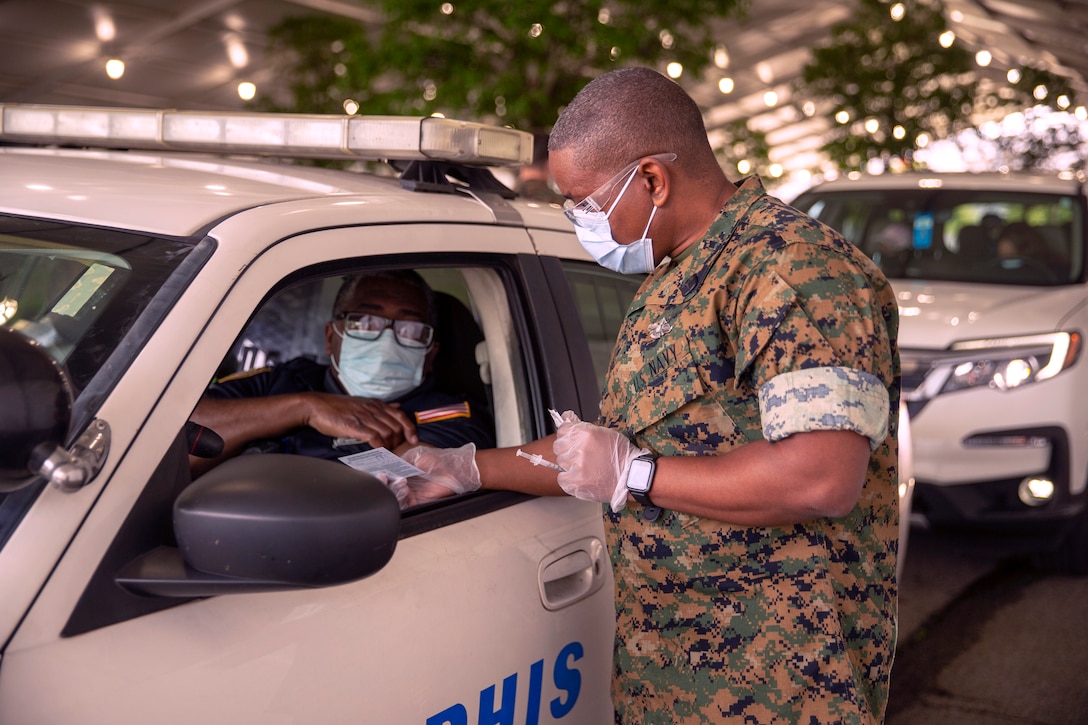 A sailor wearing a face mask and gloves speaks to a resident wearing a face mask and seated in a vehicle.