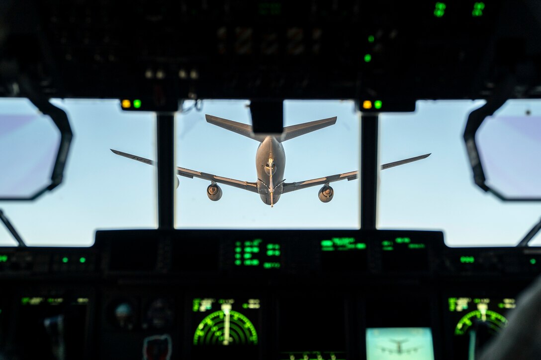 An aircraft flies in front of another aircraft as seen from a cockpit illuminated by green lights.