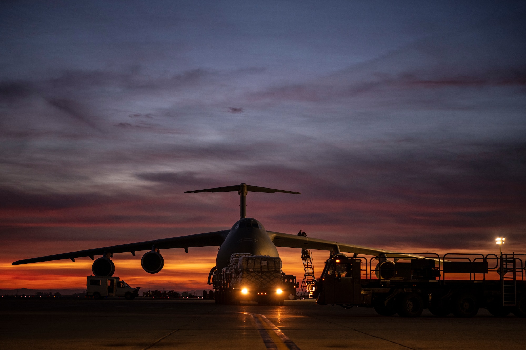 A large military vehicle is seen carrying a bunch of pallets and a colorful sky lights up behind it.