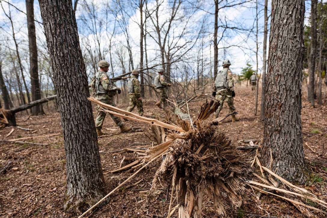 Soldiers walk through a wooded area.