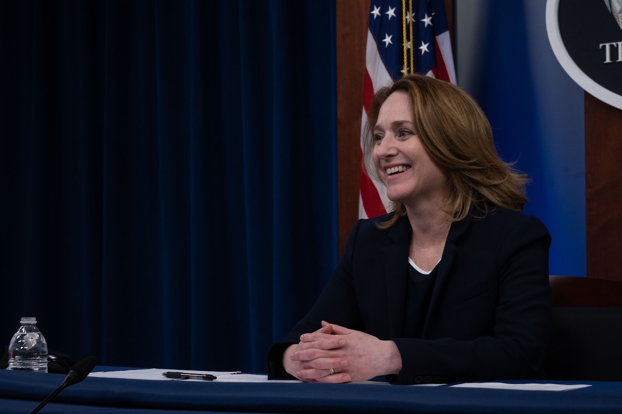 A woman speaks while seated at a table with an American flag behind her.