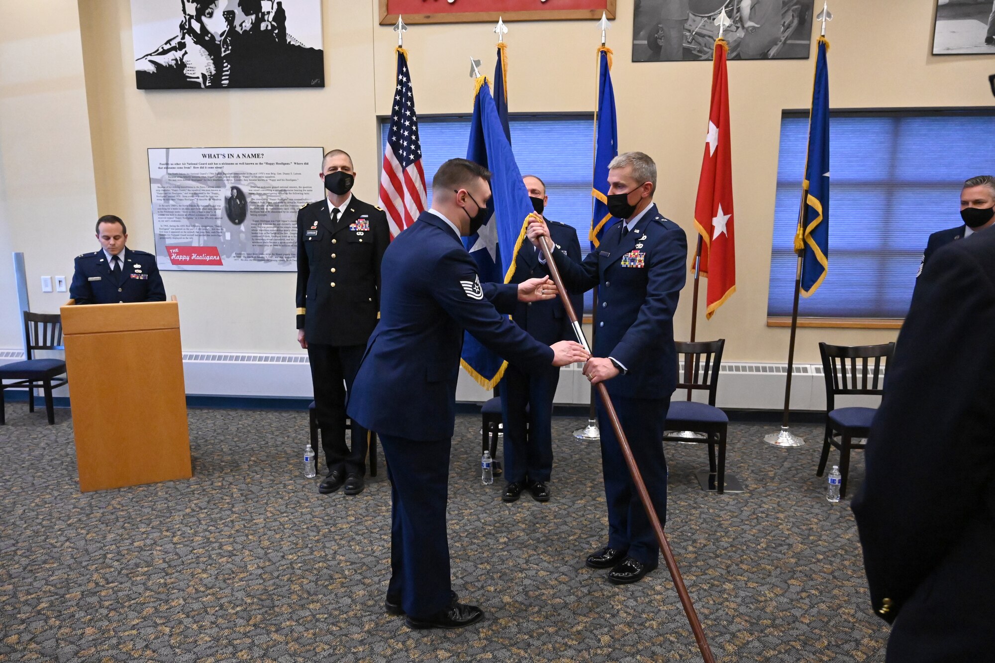 Brig. Gen. Darrin Anderson accepts a symbolic flag staff representing his assumption of responsibility for the position of North Dakota assistant adjutant general for Air from another Airman who is handing him the flag staff during a formal ceremony at the North Dakota Air National Guard Base, Fargo, N.D., April 10, 2021. A party of dignitaries watch the ceremony as they stand behind him.