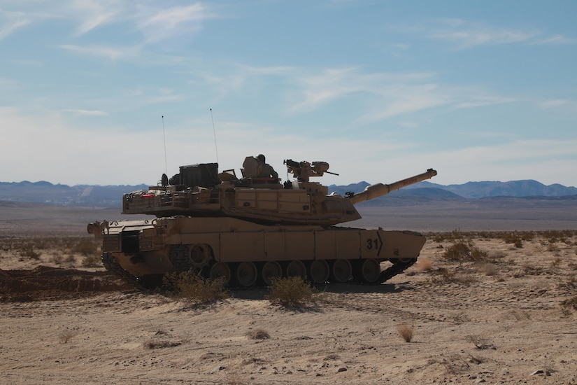 A tank sits in a dusty, desert landscape with mountains in the background.