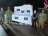 Three men in military uniforms and one in civilian clothes pose in front of a shipment of supplies covered with a protective sheet adorned with the flags of the U.S. and Paraguay.