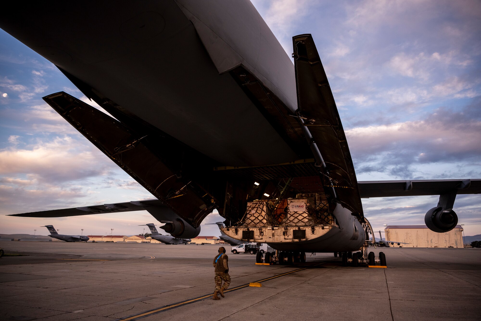 An Airmen walks towards the back of a large grey military aircraft full of pallets with USAID signs on them.