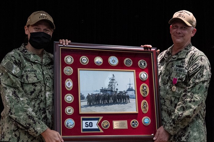 210729-N- IG466-0092 NAVAL SUPPORT ACTIVITY BAHRAIN (July 29, 2021) – Lt. Tabitha McAdams, left, presents Capt. Christopher Gilbertson, outgoing commander of Destroyer Squadron (DESRON) 50 and Task Force (TF) 55, with a farewell gift during a change of command ceremony presided over by Vice Adm. Brad Cooper, commander, U.S. Naval Forces Central Command, U.S. 5th Fleet and Combined Maritime Forces, onboard Naval Support Activity Bahrain, July 29. DESRON 50 and TF 55 operate in the U.S. 5th Fleet area of operations in support of naval operations to ensure maritime stability and security in the Central Region, connecting the Mediterranean and Pacific through the western Indian Ocean and three critical chokepoints to the free flow of global commerce. (U.S. Navy photo by Mass Communication Specialist 2nd Class Adelola Tinubu)