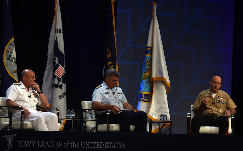 Three men in military uniforms sit in white chairs. One of them speaks.