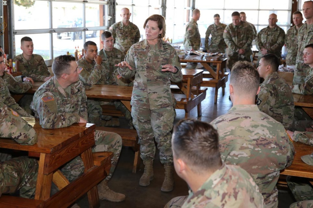A woman dressed in a military uniform stands surrounded by service members, some sitting at wooden picnic tables and some standing.