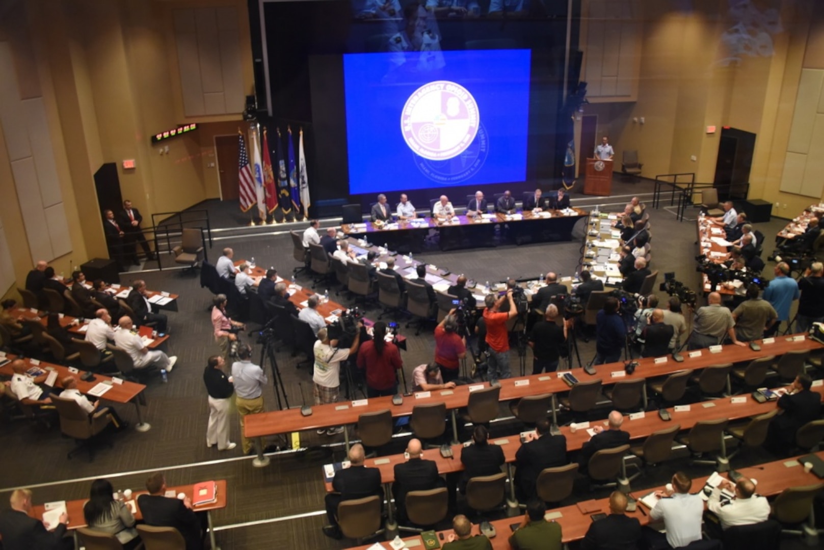 People are seated at tables in a large conference room; a large screen is in the center of the room.