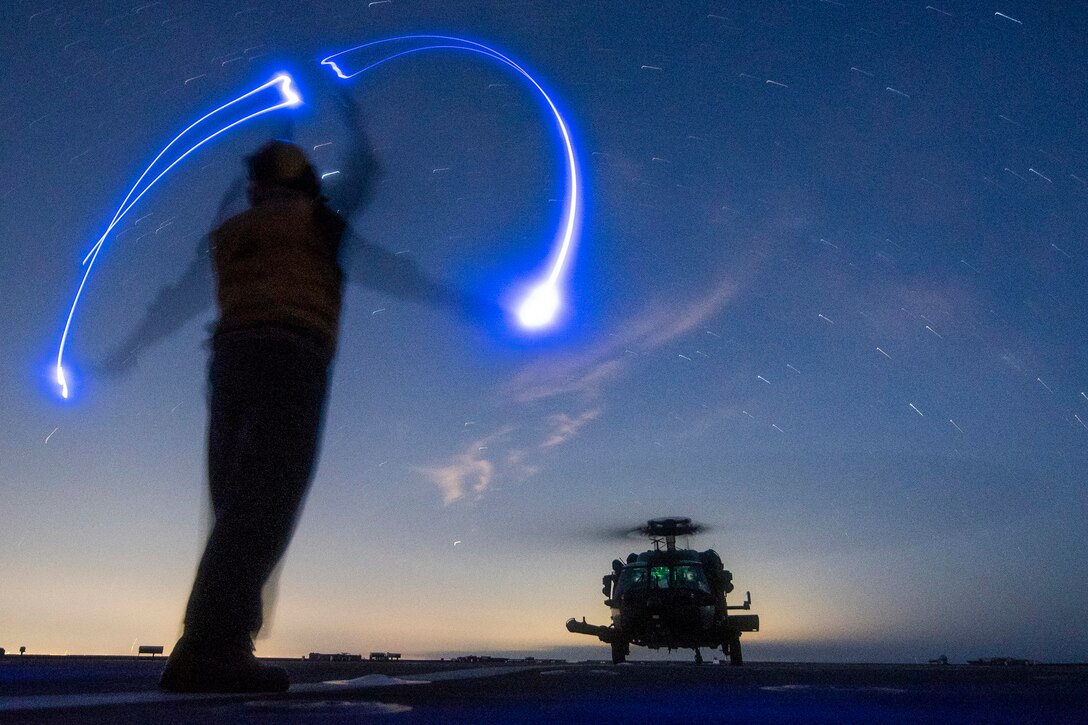 A sailor waves lights, creating an illuminated blue semicircle, as a helicopter lands.