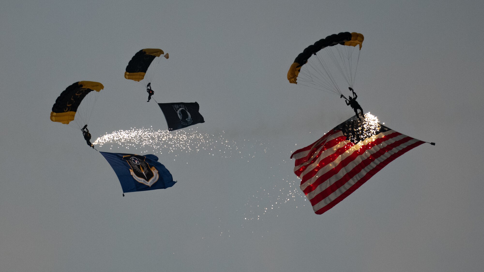 3 Special Tactics operators descend by combat parachute trailing sparklers and the United States flag along with the Air Force Special Operations Command flags.
