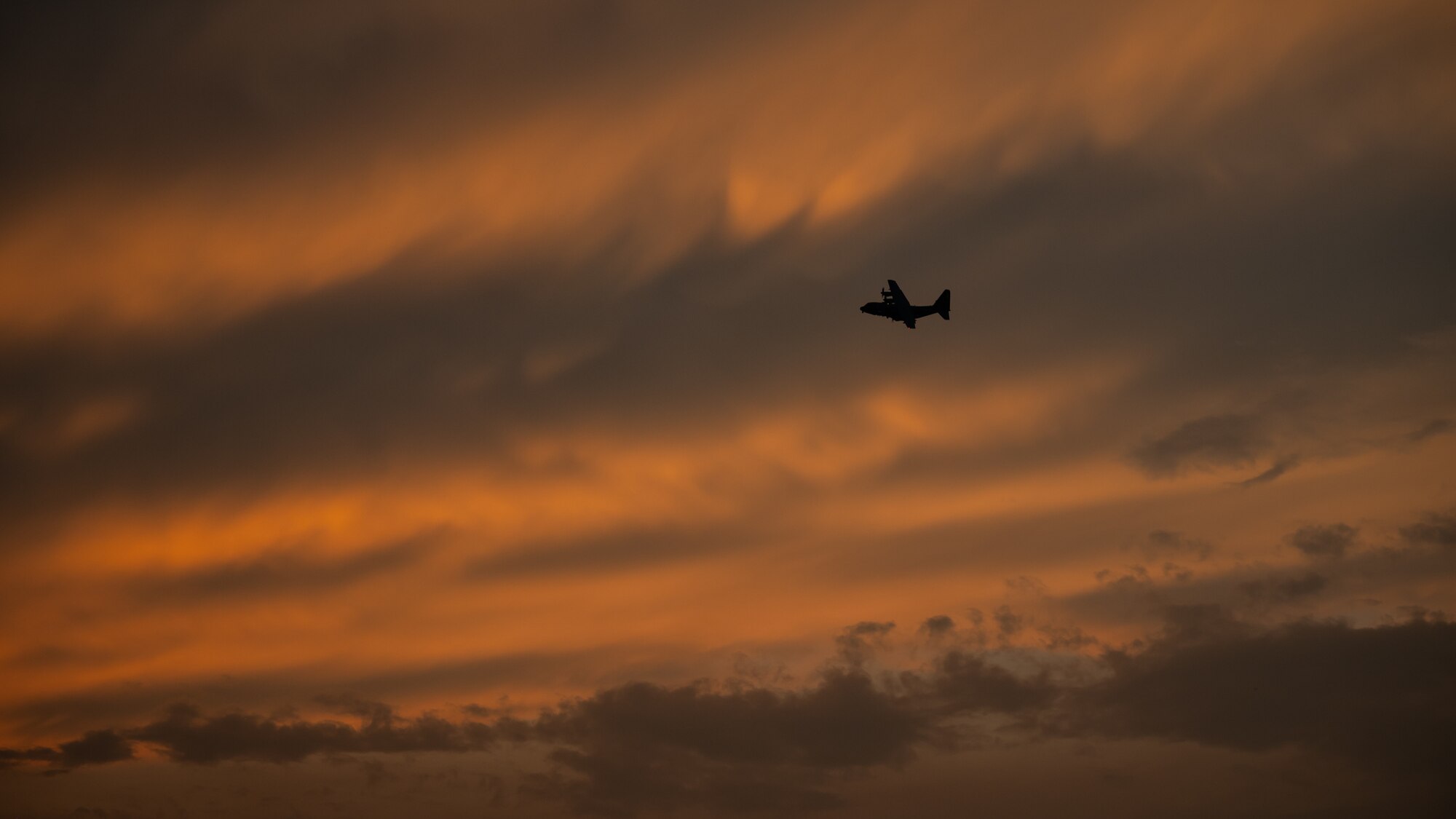 A C-130 aircraft flies in front of orange clouds during sunset.