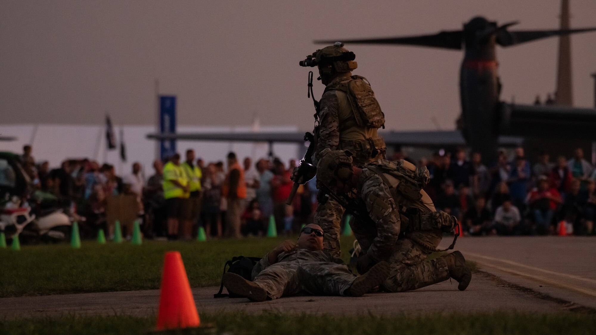 Special Tactics operators simulate rescuing a person in front of a crowd in the distance. The crowd is standing in front of a large V-22 Osprey tilt-rotor aircraft.