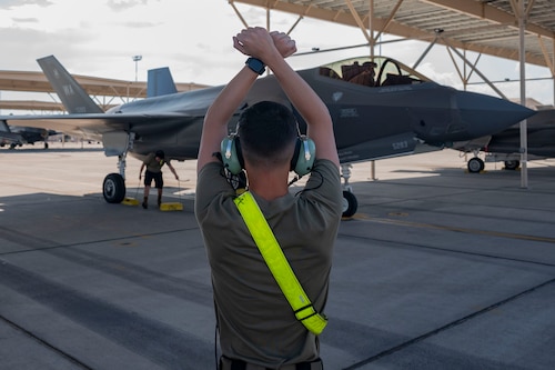 crew chief with hands in air with wrists crossed to the side of an F-35 on the flight line