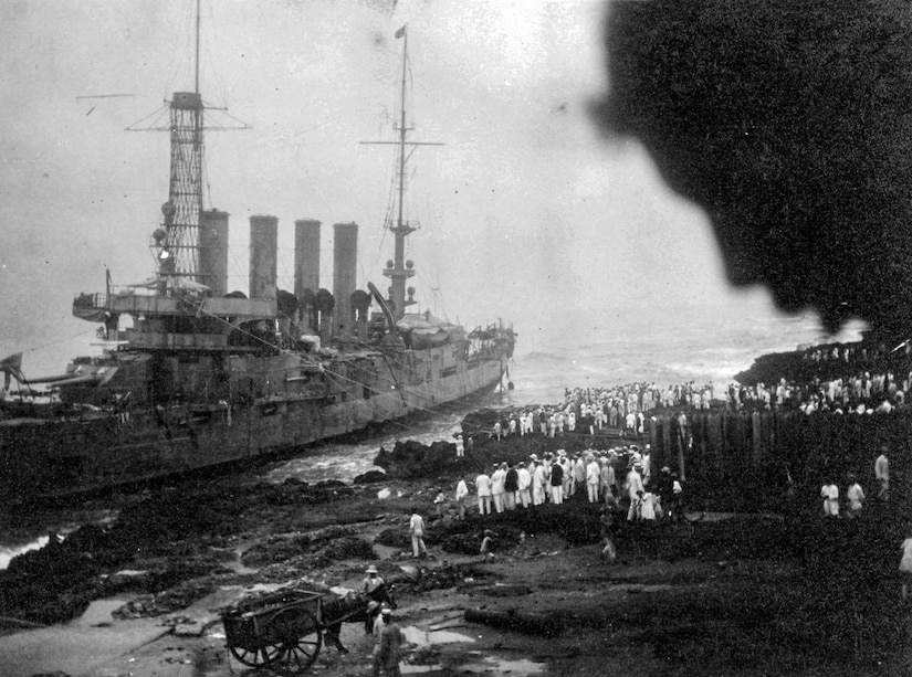 People along a shoreline stare at a large ship leaning to one side.