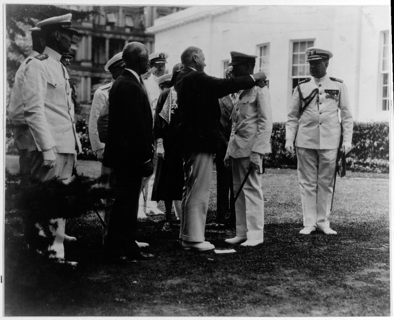 A man puts a medal around the neck of a sailor in dress whites as several other men and a woman stand around them.