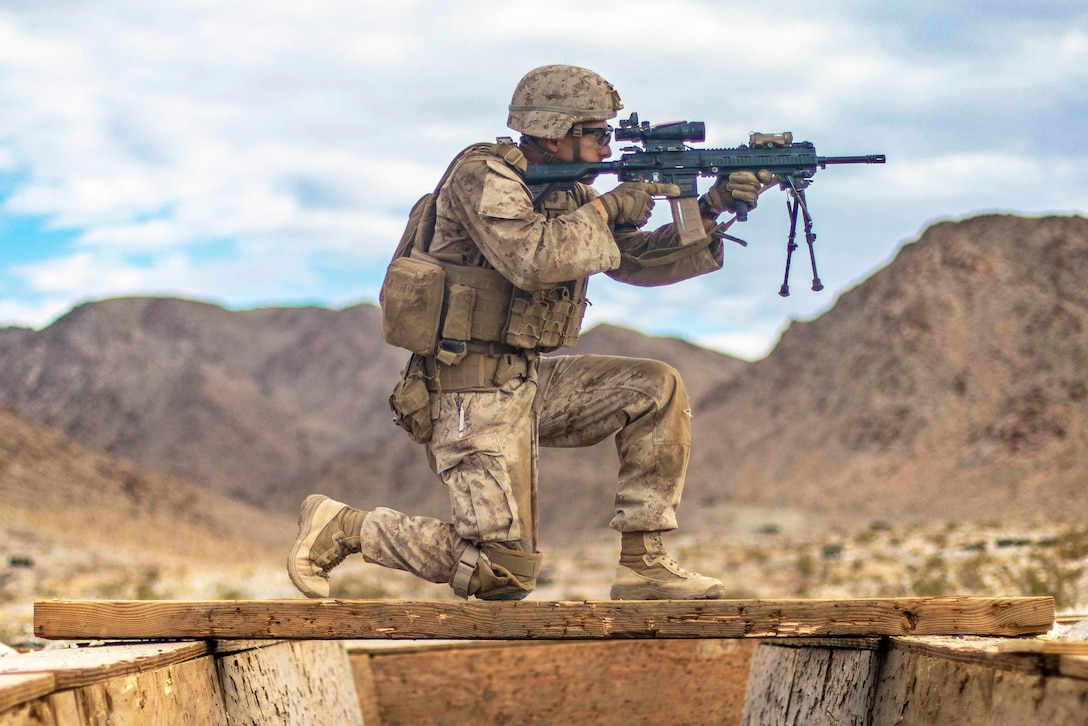 A Marine kneels on a wooden plank with an aimed weapon.