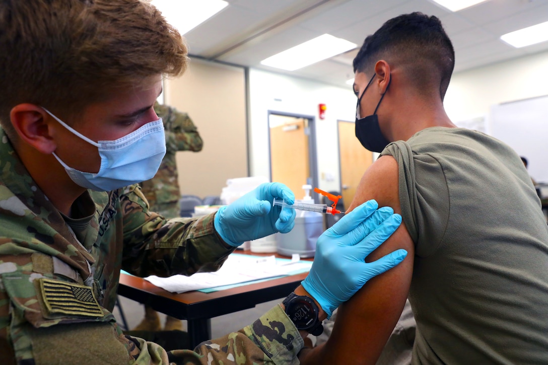 A soldier wearing a face mask and gloves leans forward to give an injection to another soldier.