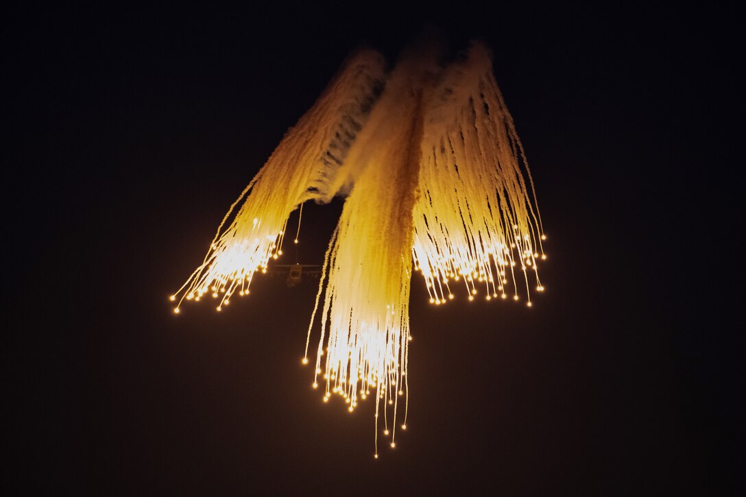 Flares descend in a night sky as an aircraft flies in the background.