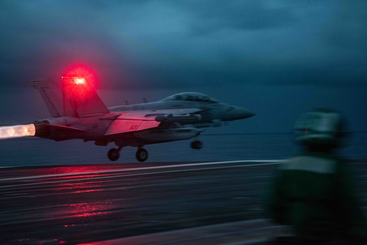 A military jet leaves the deck of a ship. It is night and the red taillight of the airplane can be seen, as can the flame from its exhaust.