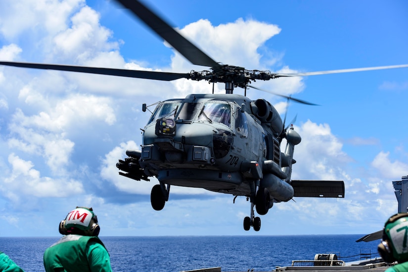 A helicopter hovers near the edge of a boat. A person in a green shirt stands nearby.