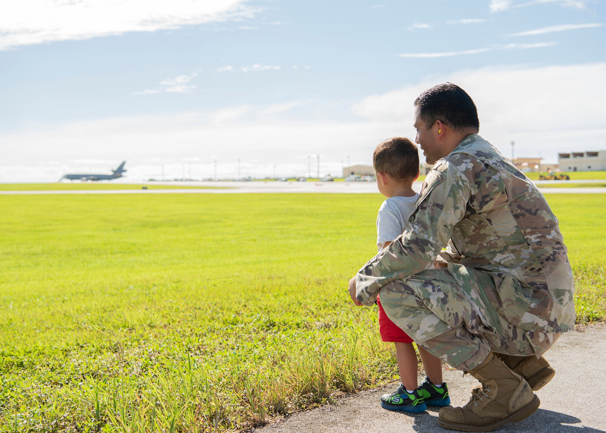A U.S. Air Force Airman watches planes take off with his son during a flight line engagement for Pacific Iron 2021 at Andersen Air Force Base, Guam, Aug. 4, 2021. PACIRON provides effective, flexible, and capabilities-centered forces, ready for deployment worldwide, and enables real-world proficiency in response to a crisis. (U.S. Air Force photo by Senior Airman Aubree Owens)